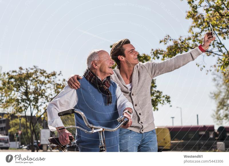 Älterer Mann und erwachsener Enkel machen ein Selfie im Freien Spaß Spass Späße spassig Spässe spaßig lächeln Handy Mobiltelefon Handies Handys Mobiltelefone