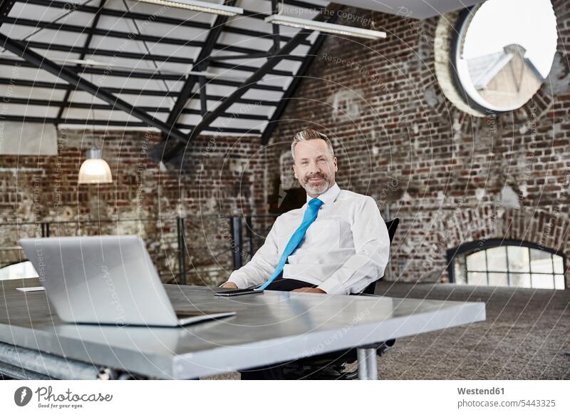 Porträt eines selbstbewussten Geschäftsmannes mit Laptop, der in einem Loft am Tisch sitzt Businessmann Businessmänner Geschäftsmänner sitzen sitzend Notebook