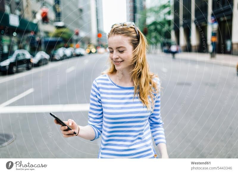 USA, New York, Manhattan, Junge Frau, die auf der Straße geht und ein Mobiltelefon in der Hand hält Handy Handies Handys Mobiltelefone Nachricht Mitteilung