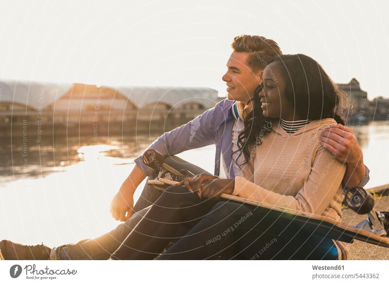 Junges Paar sitzt am Fluss und betrachtet den Sonnenuntergang multikulturell beobachten zuschauen ansehen Pärchen Paare Partnerschaft umarmen Umarmung