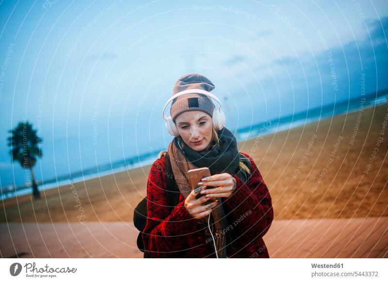 Junge Frau, die in der Dämmerung am Strand mit einem Smartphone Musik hört Handy Mobiltelefon Handies Handys Mobiltelefone weiblich Frauen Kopfhörer Kopfhoerer