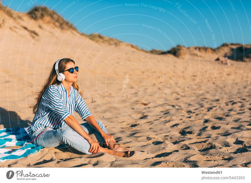 Portugal, Aveiro, Frau sitzt in der Nähe einer Stranddüne und hört Musik mit Kopfhörern Strandduene Strandduenen Stranddünen sitzen sitzend weiblich Frauen Sand