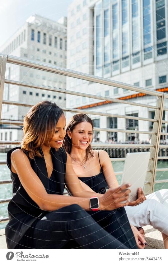 Zwei glückliche Frauen sitzen auf einer Brücke und teilen sich eine Tafel in der Stadt Kollegin Kolleginnen lächeln weiblich Tablet Tablet Computer Tablet-PC