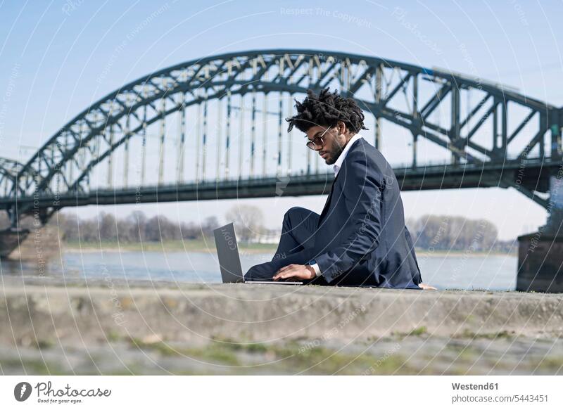 Geschäftsmann sitzt mit Laptop am Flussufer Businessmann Businessmänner Geschäftsmänner Notebook Laptops Notebooks Mann Männer männlich Geschäftsleute