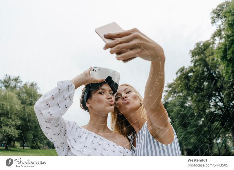 Zwei junge Frauen mit VR-Brille machen ein Selfie in einem Park Parkanlagen Parks Brillen Freundinnen Selfies weiblich Virtuelle Realität Virtuelle Realitaet