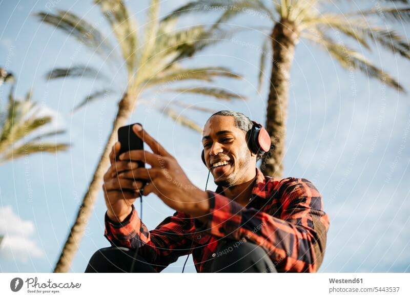 Spanien, Porträt eines lächelnden jungen Mannes, der mit Kopfhörern Musik hört und auf sein Handy schaut Kopfhoerer Männer männlich Erwachsener erwachsen Mensch