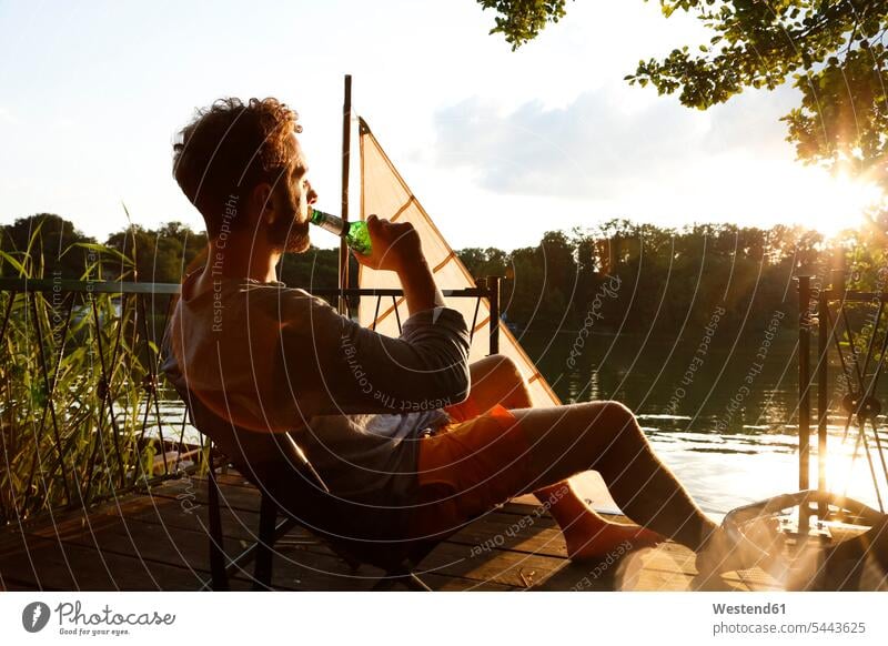 Junger Mann mit Biertrinken auf einem Steg neben einem Segelboot Stege Anlegestelle Männer männlich Segelboote Segelschiff sitzen sitzend sitzt Alkohol