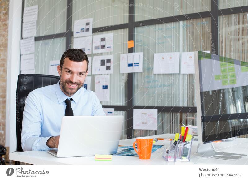 Porträt eines lächelnden Geschäftsmannes im Büro mit Laptop am Schreibtisch Businessmann Businessmänner Geschäftsmänner Notebook Laptops Notebooks