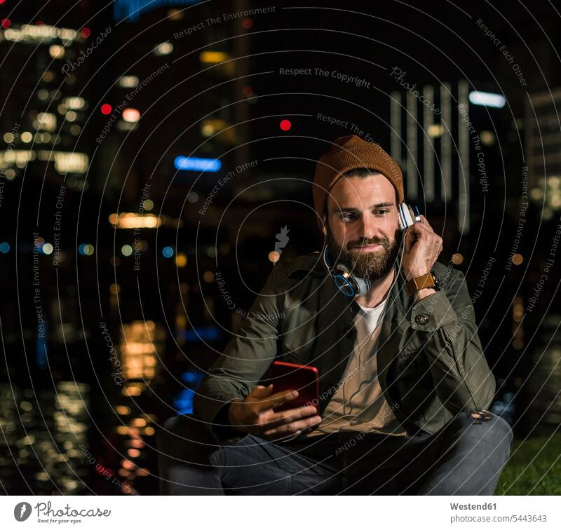 Stylischer junger Mann mit Handy und Kopfhörer sitzt nachts am städtischen Flussufer Kopfhoerer lächeln Nacht Männer männlich Mobiltelefon Handies Handys