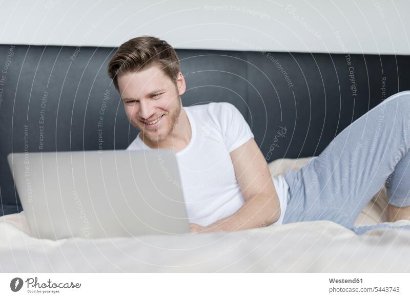 Lächelnder junger Mann liegt mit Laptop auf dem Bett Betten lächeln Notebook Laptops Notebooks Männer männlich Computer Rechner Erwachsener erwachsen Mensch