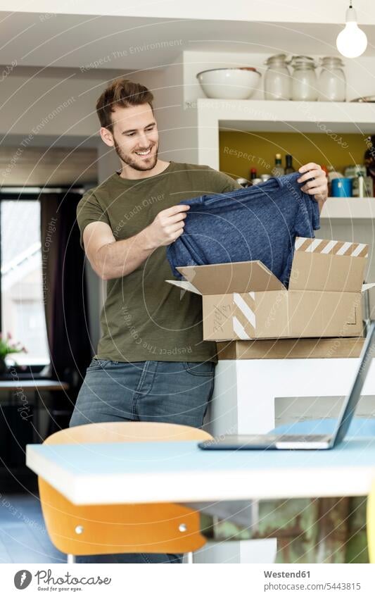Lächelnder junger Mann zu Hause, der ein Paket mit Kleidung auspackt Zuhause daheim Pakete Päckchen lächeln Kleider auspacken Männer männlich Erwachsener