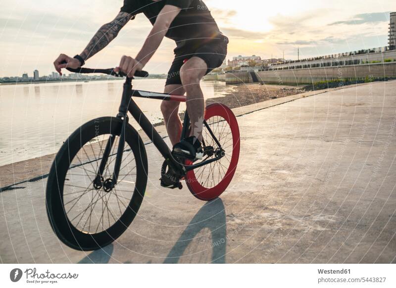 Junger Mann fährt Fixie-Rad am Wasser bei Sonnenuntergang Fahrrad Bikes Fahrräder Räder Fluss Fluesse Fluß Flüsse Männer männlich fahren fahrend fahrender