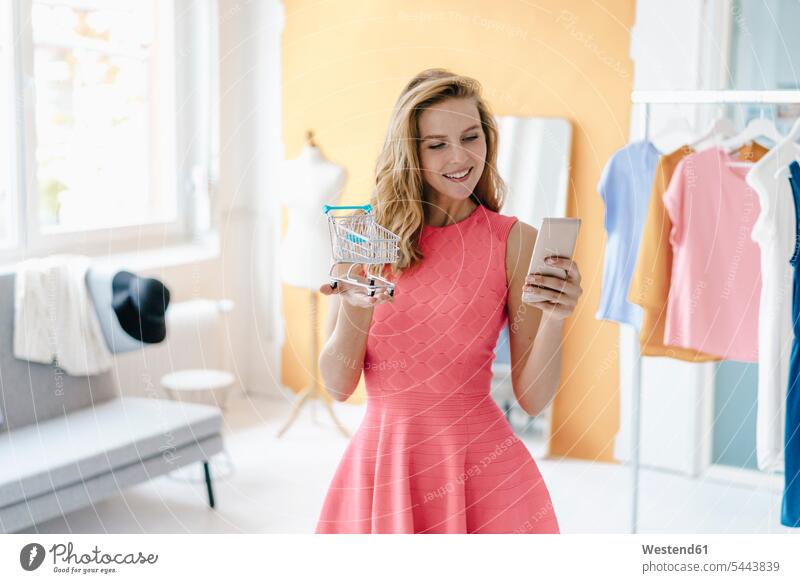 Lächelnde junge Frau im Mode-Studio beim Fotografieren eines Einkaufswagen-Models mit dem Handy lächeln Atelier Studios Ateliers Mobiltelefon Handies Handys