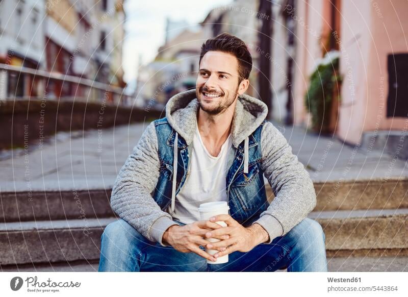 Mann mit Kaffee sitzt auf einer Treppe mit der Stadt im Hintergrund glücklich Glück glücklich sein glücklichsein sitzen sitzend Männer männlich lächeln