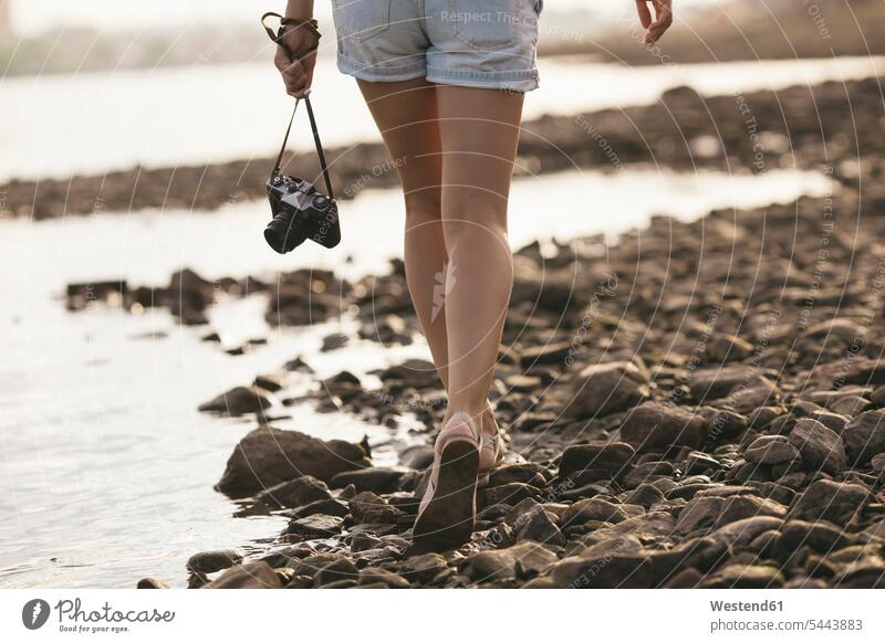 Frau mit einer Kamera in der Hand geht am steinigen Strand Fluss Fluesse Fluß Flüsse gehen gehend weiblich Frauen Fotoapparat Fotokamera Gewässer Wasser