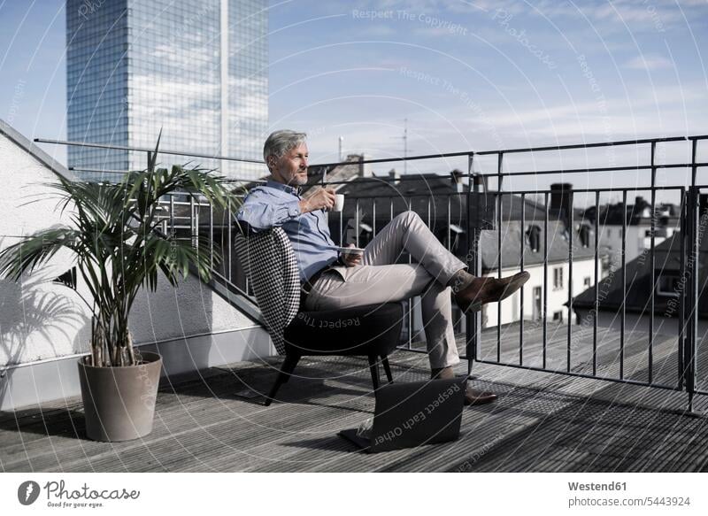 Grauhaariger Geschäftsmann sitzt auf dem Balkon mit Blick auf die Stadt und trinkt Kaffee Businessmann Businessmänner Geschäftsmänner Laptop Notebook Laptops