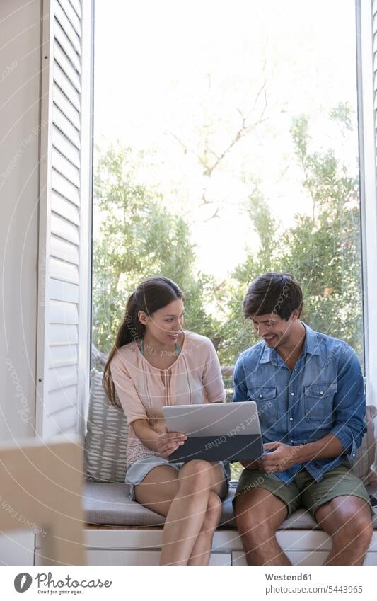 Lächelndes junges Paar sitzt auf der Fensterbank und teilt sich ein Tablett Pärchen Paare Partnerschaft entspannt entspanntheit relaxt Tablet Computer Tablet-PC