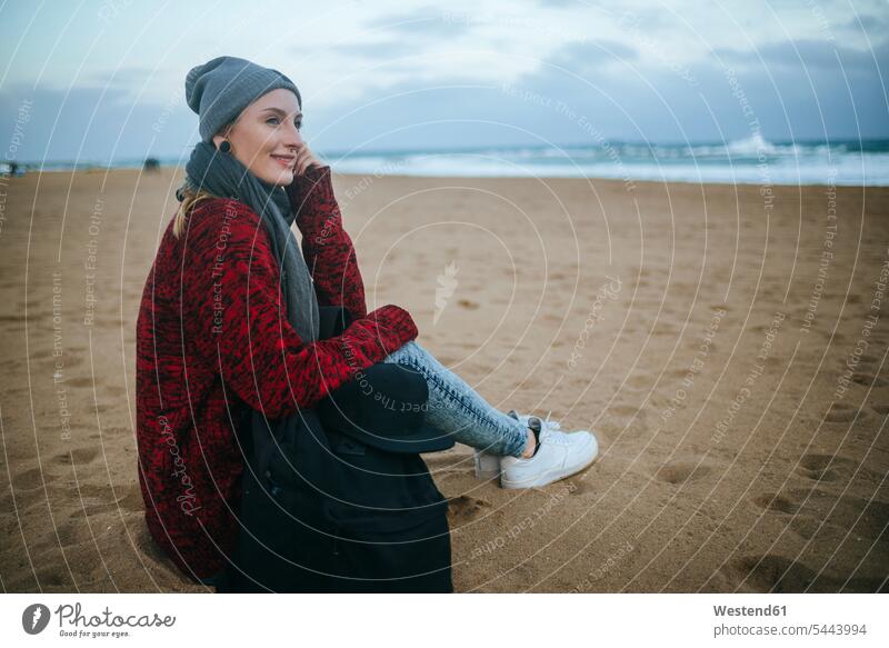 Lächelnde junge Frau sitzt im Winter am Strand Beach Straende Strände Beaches sitzen sitzend weiblich Frauen lächeln Erwachsener erwachsen Mensch Menschen Leute