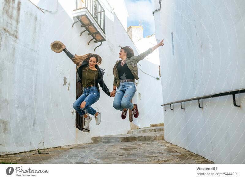 Zwei glückliche junge Frauen springen in einer Gasse einer Stadt Spaß Spass Späße spassig Spässe spaßig Freundinnen hüpfen Freunde Freundschaft Kameradschaft