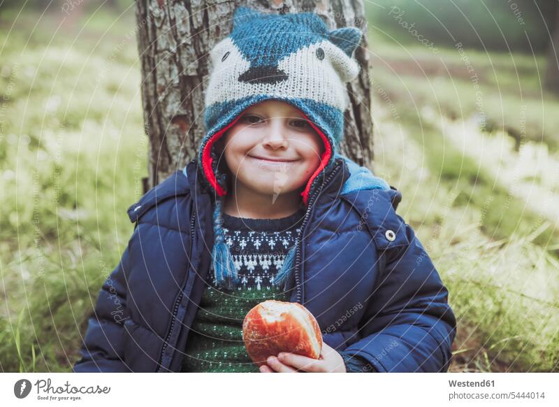Porträt eines grinsenden Jungen, der im Wald einen Berliner isst essen essend Buben Knabe Knaben männlich Wollmütze Wollmützen Strickmütze Strickmützen Kind