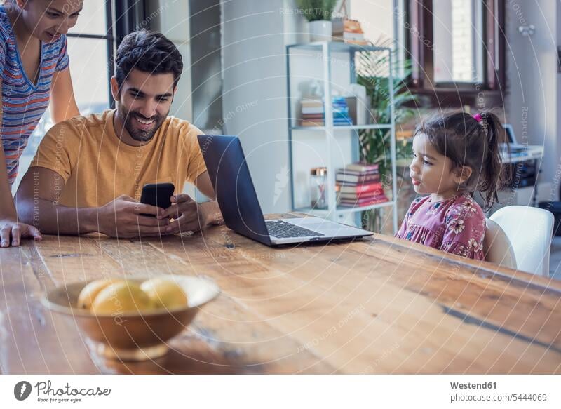 Familie sitzt in der Küche, Eltern benutzen ein Smartphone, die Tochter schaut auf den Laptop Familien benützen iPhone Smartphones Notebook Laptops Notebooks