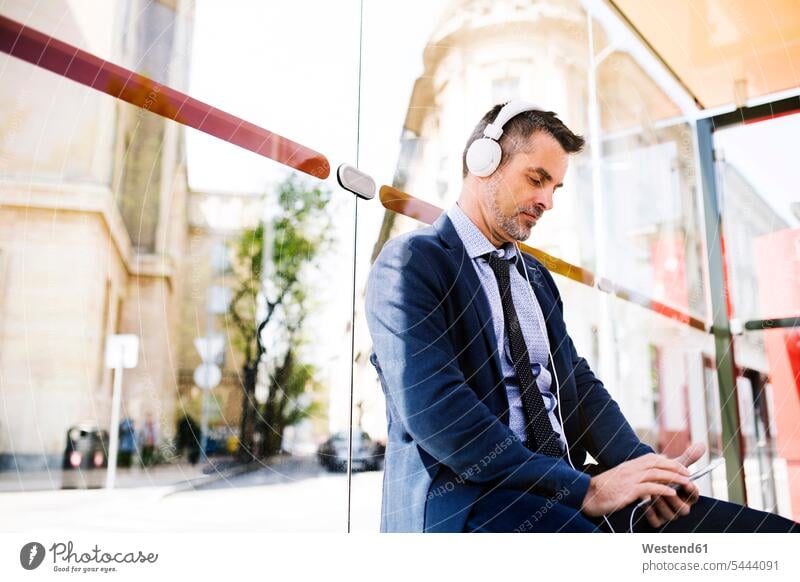 Geschäftsmann mit Smartphone und Kopfhörern wartet an der Bushaltestelle Handy Mobiltelefon Handies Handys Mobiltelefone Pendler Haltestelle Haltestellen