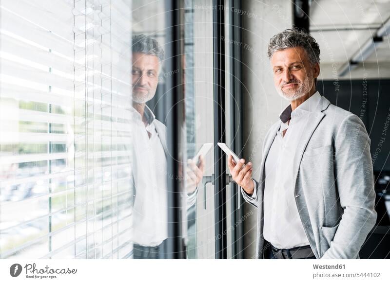 Porträt eines lächelnden reifen Geschäftsmannes am Fenster im Büro mit einem Mobiltelefon in der Hand Office Büros halten Handy Handies Handys Mobiltelefone