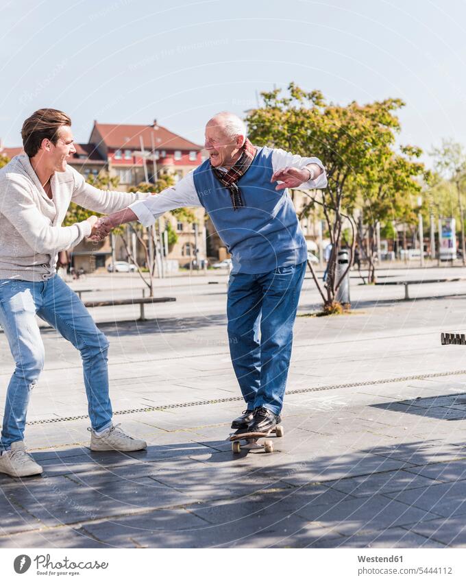 Erwachsener Enkel unterstützt älteren Mann auf Skateboard Rollbretter Skateboards Großvater Opa Großpapa Großpapas Opas Opi Großväter Opis glücklich Glück