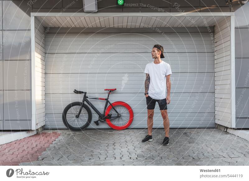 Junger Mann mit Fixie-Rad an einem Rollladen stehen stehend steht Fahrrad Bikes Fahrräder Räder Männer männlich Raeder Erwachsener erwachsen Mensch Menschen