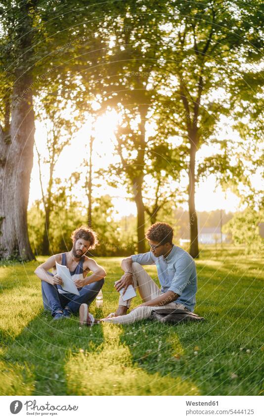 Zwei Freunde sitzen in einem Park mit mobilem Gerät und Papieren Mann Männer männlich Parkanlagen Parks Handy Mobiltelefon Handies Handys Mobiltelefone