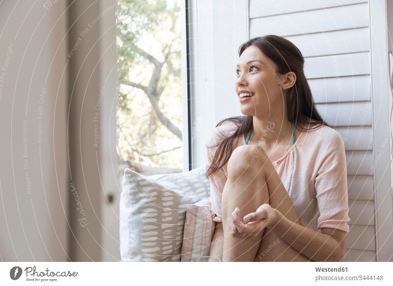 Lächelnde junge Frau sitzt auf der Fensterbank entspannt entspanntheit relaxt sitzen sitzend weiblich Frauen lächeln Entspannung relaxen entspannen Erwachsener