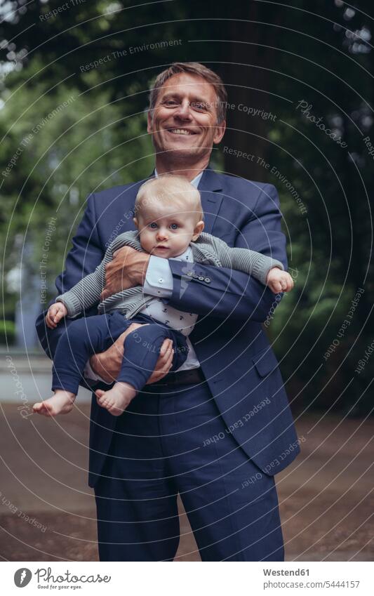 Porträt eines lächelnden, reifen Geschäftsmannes, der einen kleinen Jungen im Freien hält Baby Babies Babys Säuglinge Kind Kinder Vater Papas Väter Vati Vatis