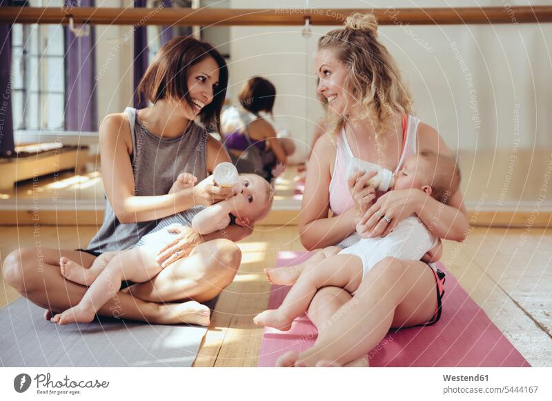 Zwei Mütter füttern ihre Babys im Übungsraum mit der Flasche Mutter Mami Mutti Mama Spaß Spass Späße spassig Spässe spaßig trainieren lächeln Babies Säuglinge