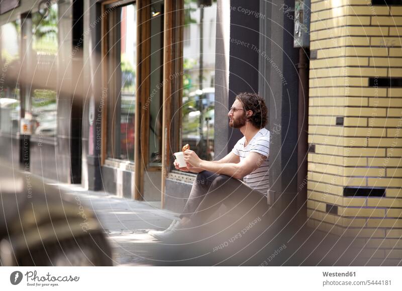 Junger Mann mit Kaffeetasse und Croissant am Eingang eines Cafés sitzend Männer männlich Erwachsener erwachsen Mensch Menschen Leute People Personen Eingänge