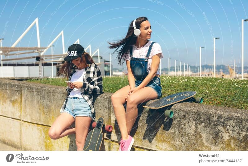 Junge Frau mit Kopfhörern hört Musik, während ihre Freundin ein Smartphone anschaut Longboard Freundinnen Skateboarderin Skateboardfahrerin Skaterin