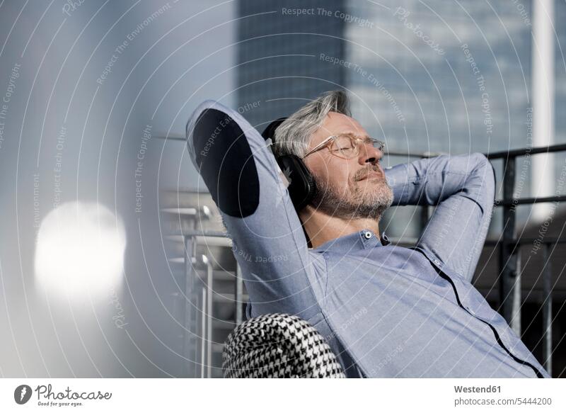 Grauhaariger Mann entspannt sich in einem Stuhl auf dem Balkon und hört Musik Geschäftsmann Businessmann Businessmänner Geschäftsmänner sitzen sitzend sitzt