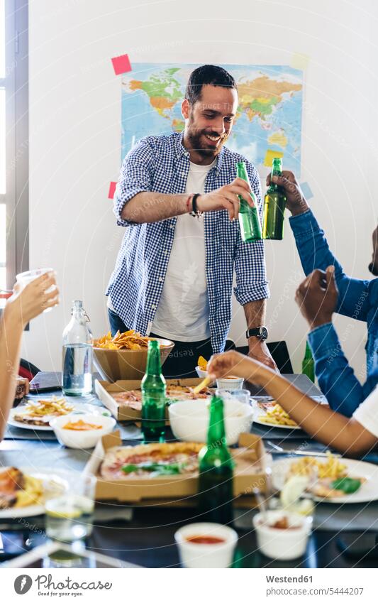 Gruppe von Freunden klirrt mit Bierflaschen am Esstisch Pizza Pizzen essen essend anstoßen zuprosten anstossen Tisch Tische trinken Alkohol