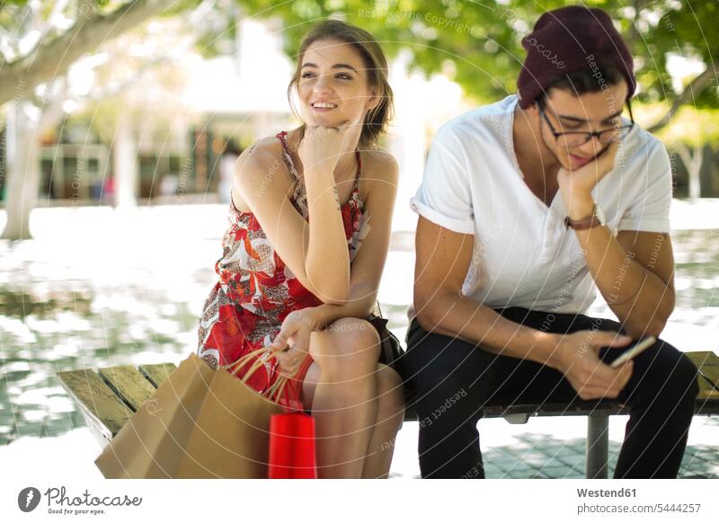 Junges Paar sitzt auf Bank mit Einkaufstaschen und Handy Shopping einkaufen shoppen Mobiltelefon Handies Handys Mobiltelefone Pärchen Paare Partnerschaft Tasche