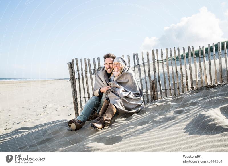 Lächelndes, in eine Decke gehülltes Paar am Strand Beach Straende Strände Beaches Pärchen Paare Partnerschaft lächeln Mensch Menschen Leute People Personen