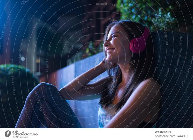 Glückliche junge Frau mit rosa Kopfhörern, die nachts in moderner städtischer Umgebung Musik hört trainieren telefonieren anrufen Anruf telephonieren