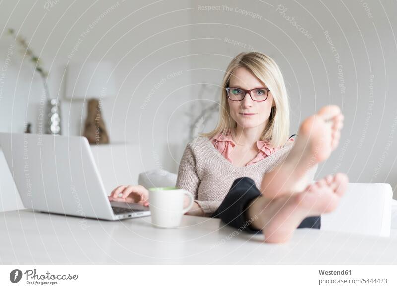 Porträt einer blonden Frau zu Hause, die mit erhobenen Füßen am Tisch sitzt Portrait Porträts Portraits Laptop Notebook Laptops Notebooks weiblich Frauen