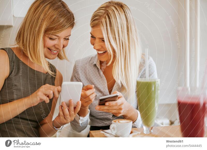 Zwei glückliche junge Frauen mit Smartphones in einem Cafe sprechen reden Freundinnen Handy Mobiltelefon Handies Handys Mobiltelefone Kaffeehaus Bistro Cafes