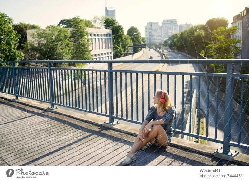 Junge Frau sitzt auf der Brücke und hört Musik mit Kopfhörern sitzen sitzend weiblich Frauen hören hoeren Kopfhoerer Bruecken Brücken lächeln Erwachsener