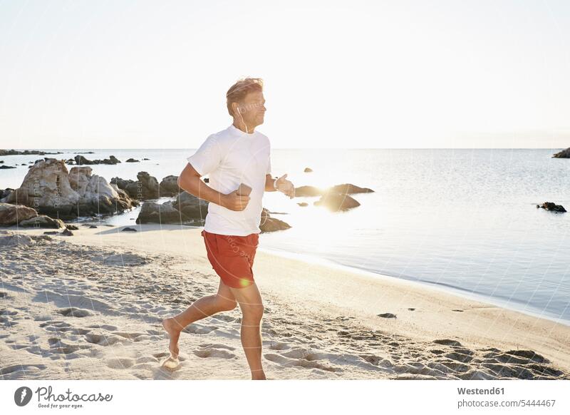 Erwachsener Mann joggt am Strand, während er mit Smartphone und Kopfhörern Musik hört Beach Straende Strände Beaches Jogger Joggen Jogging Fitness fit