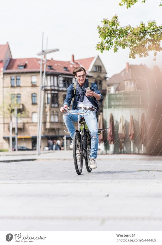 Lächelnder junger Mann mit Fahrrad in der Stadt, der auf sein Handy schaut Männer männlich Bikes Fahrräder Räder Rad lächeln Mobiltelefon Handies Handys
