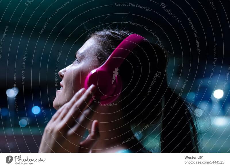 Glückliche junge Frau mit rosa Kopfhörern, die nachts in moderner städtischer Umgebung Musik hört trainieren junge Frauen telefonieren anrufen Anruf