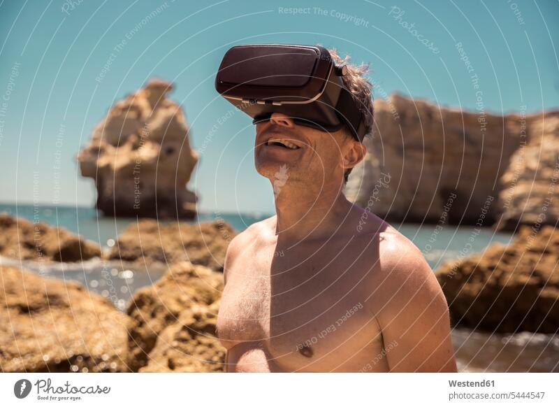 Erwachsener Mann mit VR-Brille zwischen Felsen am Strand Virtuelle Realität Virtuelle Realitaet Männer männlich Beach Straende Strände Beaches Brillen erwachsen