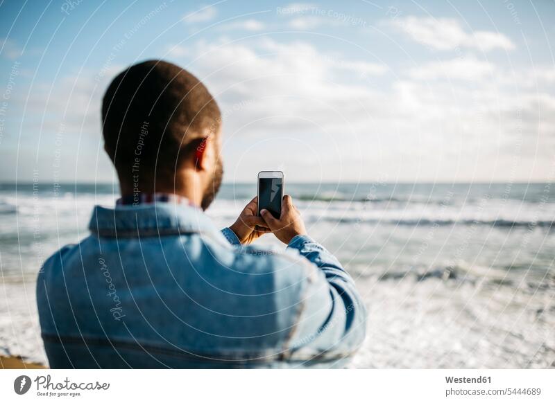 Rückenansicht eines jungen Mannes, der das Meer fotografiert Smartphone iPhone Smartphones fotografieren Handy Mobiltelefon Handies Handys Mobiltelefone Meere