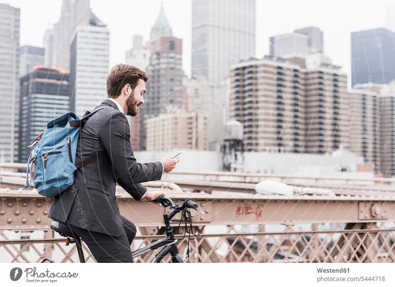 USA, New York City, Geschäftsmann auf dem Fahrrad auf der Brooklyn Bridge mit Mobiltelefon Businessmann Businessmänner Geschäftsmänner Brücke Bruecken Brücken