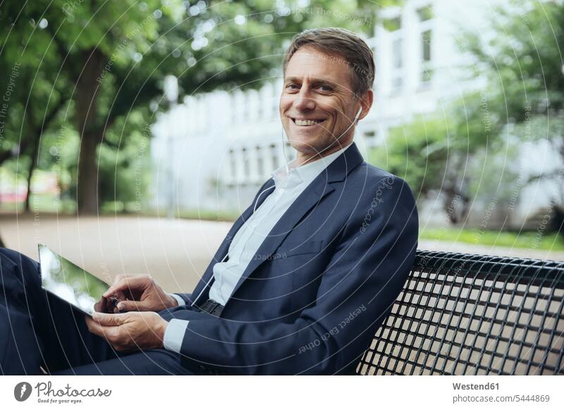 Lächelnder reifer Geschäftsmann auf Parkbank sitzend mit Tablett und Kopfhörern Ohrhörer lächeln Businessmann Businessmänner Geschäftsmänner Portrait Porträts